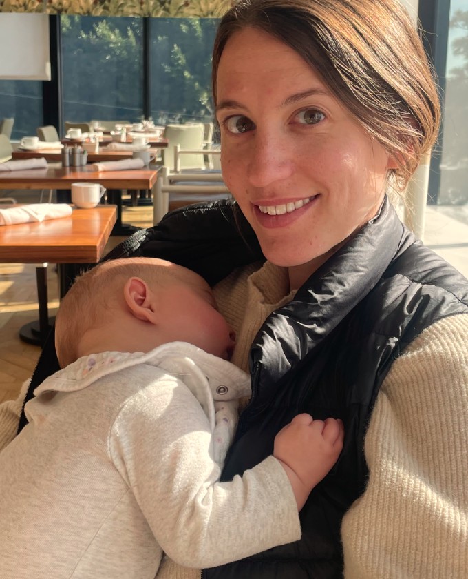 Kim smiling in a sunlit restaurant with her sleeping baby held against her chest
